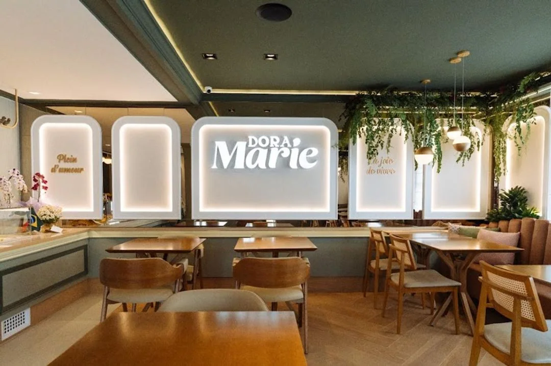 Ambiente da Dora Marie, com mesas de madeira, plantas e a marca ao fundo